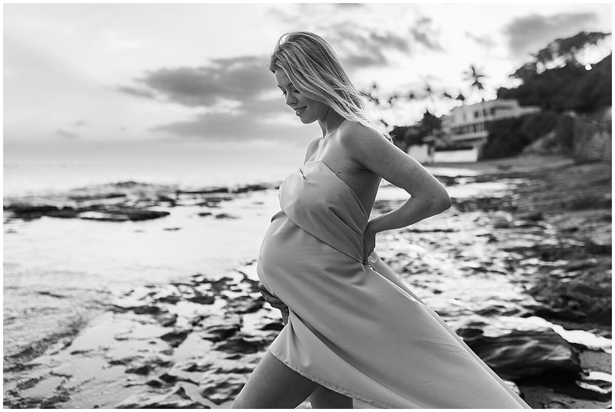 New mom holding stomach over long white gown at sunset on the beach with Elle Rose photo